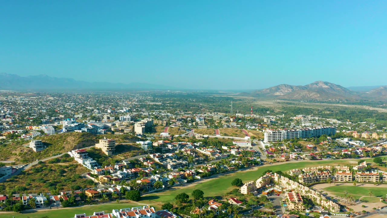 Opening aerial view over San Jose del Cabo with green space in the foreground, residential and commercial buildings across the middle distance, mountains on the horizon, and no visible people.