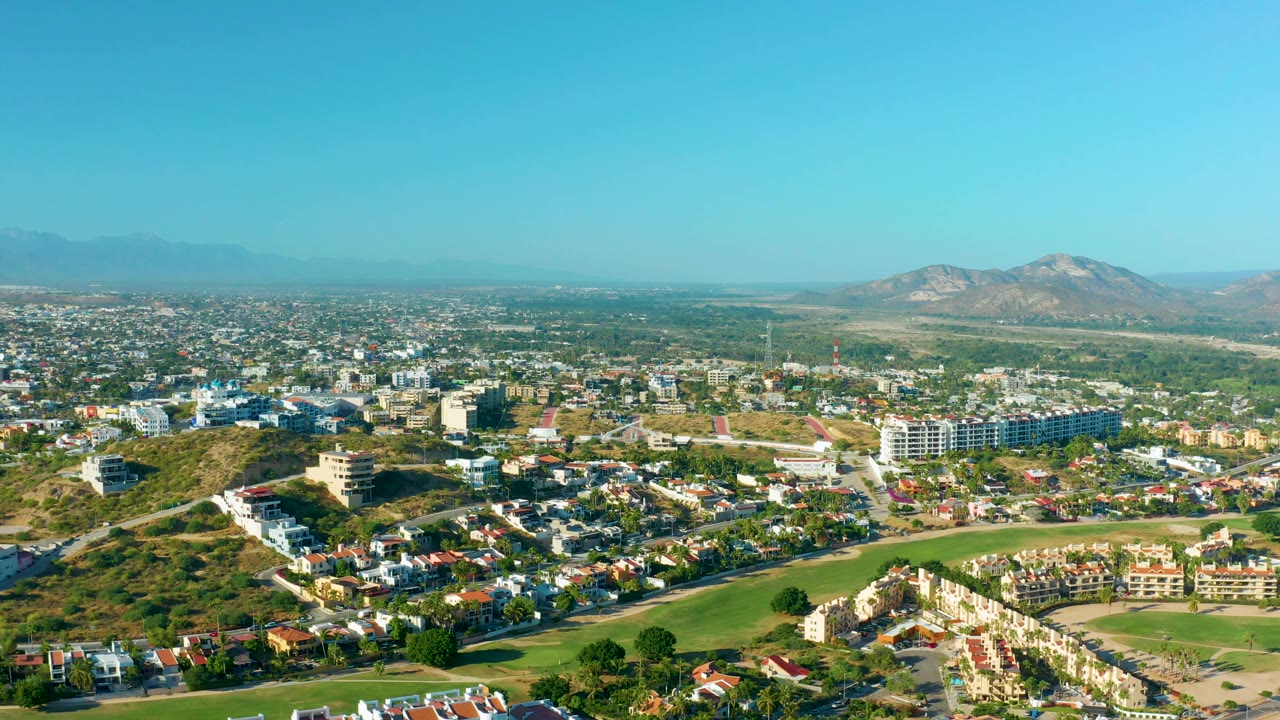 City and hillside panorama with green foreground and mountains beneath blue sky.