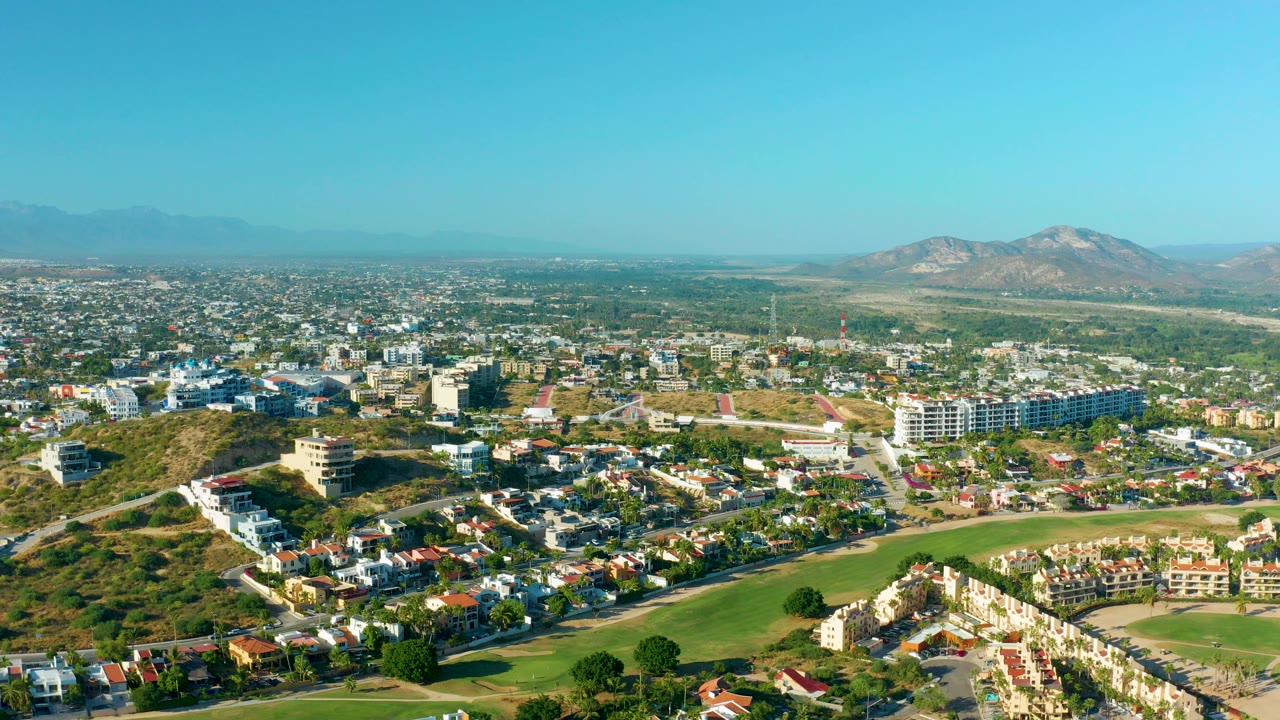 Opening aerial frame over San Jose del Cabo residential blocks, with green lawns in the foreground, hillside homes at left, distant mountains on the horizon, and no visible people.