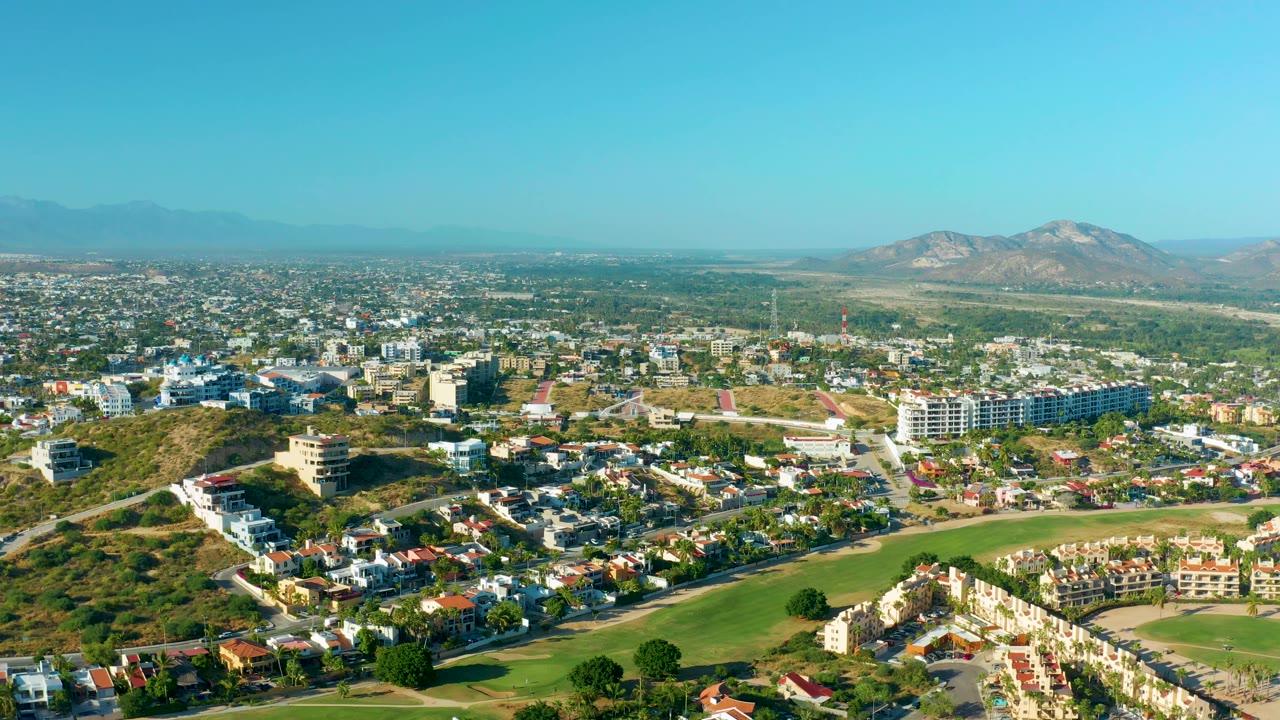 San Jose del Cabo aerial frame with dense low-rise housing, a long green lawn area near the bottom, several larger white buildings toward the right, distant mountains, and no visible people.