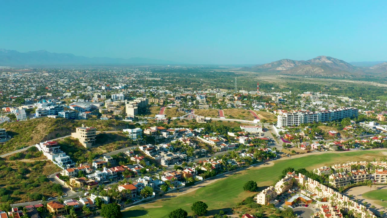 Opening aerial frame over San Jose Del Cabo, Mexico, showing residential blocks, a green field in the foreground, dry hills, distant mountains, and no visible people.