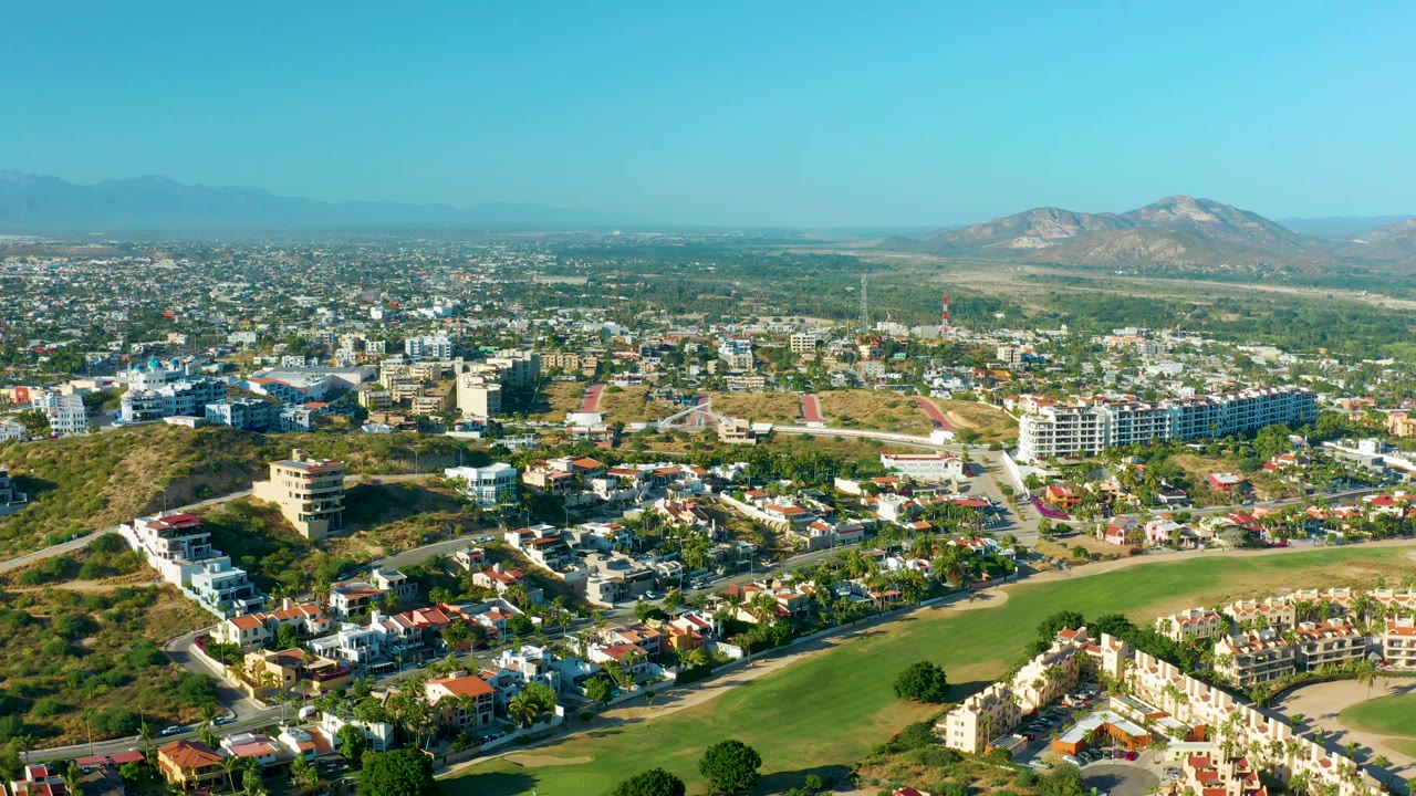 Third frame of the San Jose Del Cabo aerial view shows clustered residential buildings, a fairway-like green area in front, dry hills at left, mountains on the horizon, and no visible people.