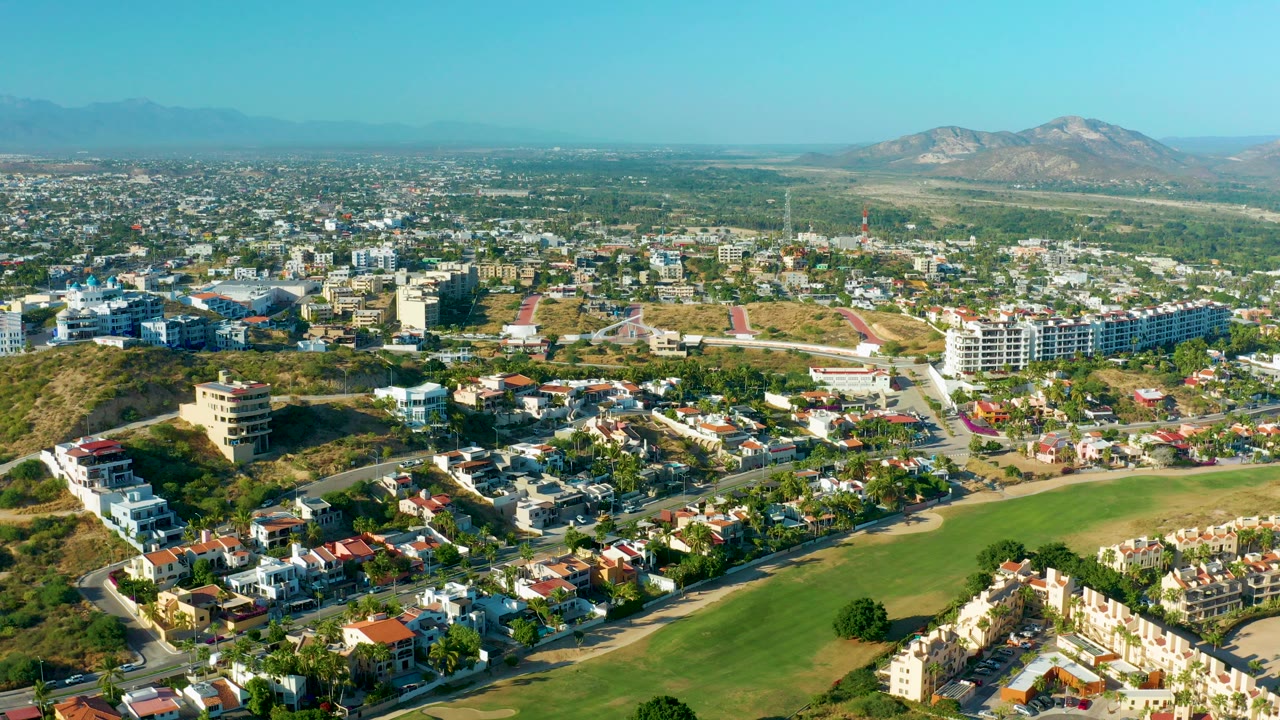 Wide aerial frame over San Jose del Cabo showing a green field in the foreground, residential blocks across the midground, dry hills, distant mountains, blue sky, and no visible people.