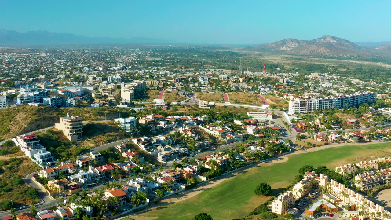 San Jose del Cabo aerial frame with clustered homes, a large green field at bottom, white and tan buildings on hillsides, distant mountains, clear blue sky, and no visible people.