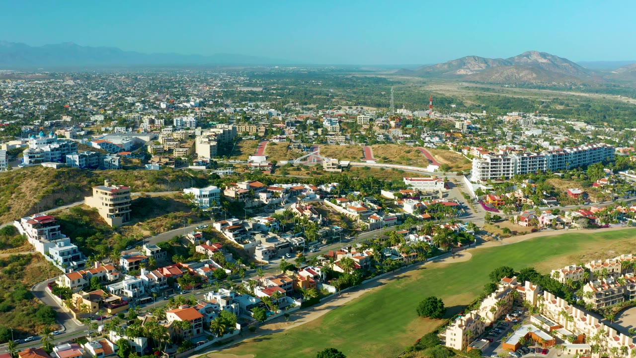 Aerial view over San Jose del Cabo with roads cutting through residential blocks, a green open field in the foreground, dry hills in the midground, distant mountains, and no visible people.