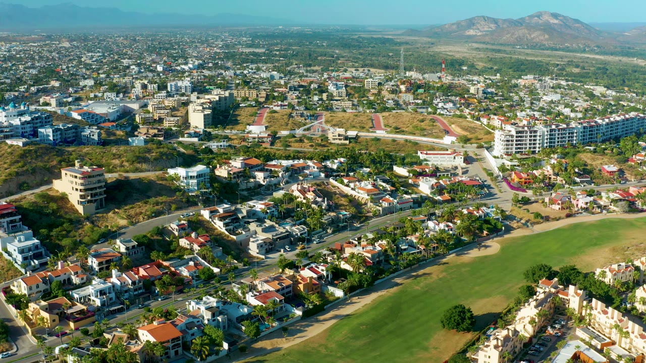 San Jose Del Cabo aerial view with the green field filling the lower right, white and red-roofed homes across the foreground, larger buildings beyond, distant mountains, and no visible people.