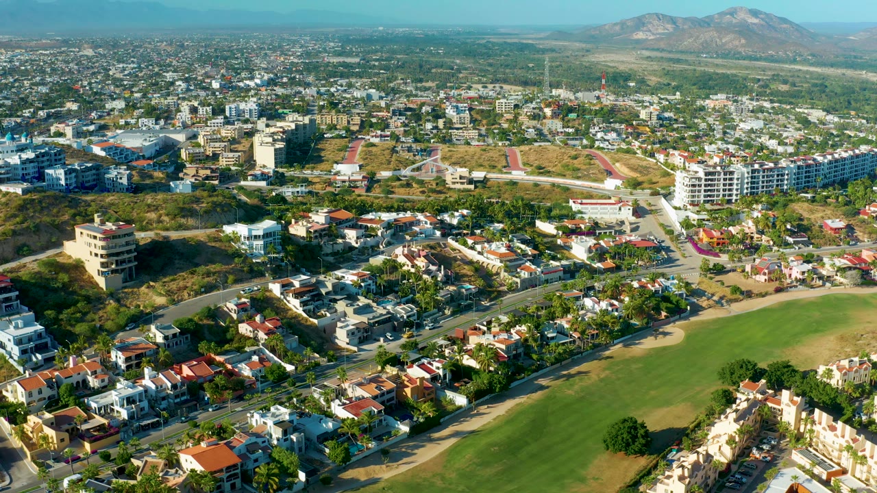 High aerial frame over San Jose Del Cabo showing a curved road beside the green field, compact residential blocks, low apartment structures, dry hills, mountains, and no visible people.