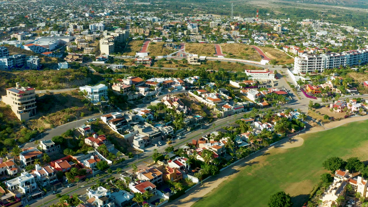 Opening aerial frame over San Jose del Cabo homes, curving roads, a large green field, sandy beach edge, and distant low-rise buildings, with no visible people.