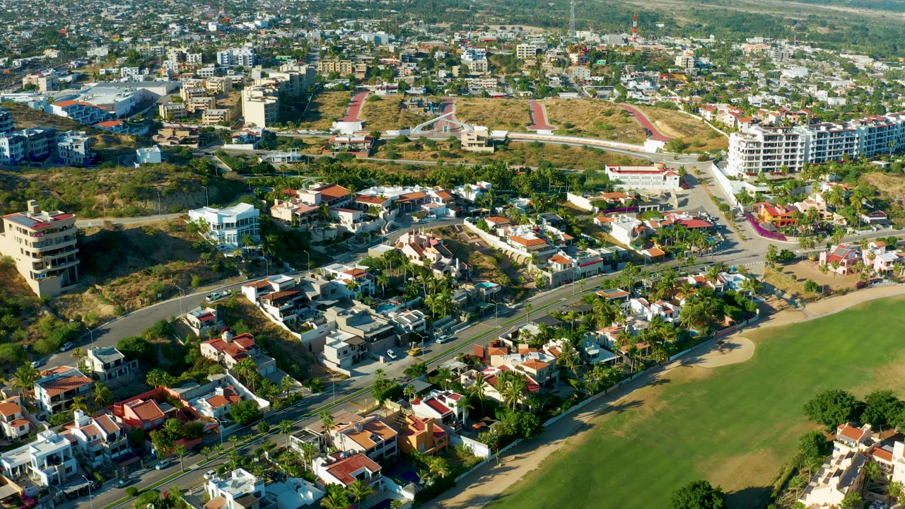 San Jose del Cabo aerial frame with low-rise apartments at the right, residential blocks below, a road junction through the center, dry open lots, and no visible people.