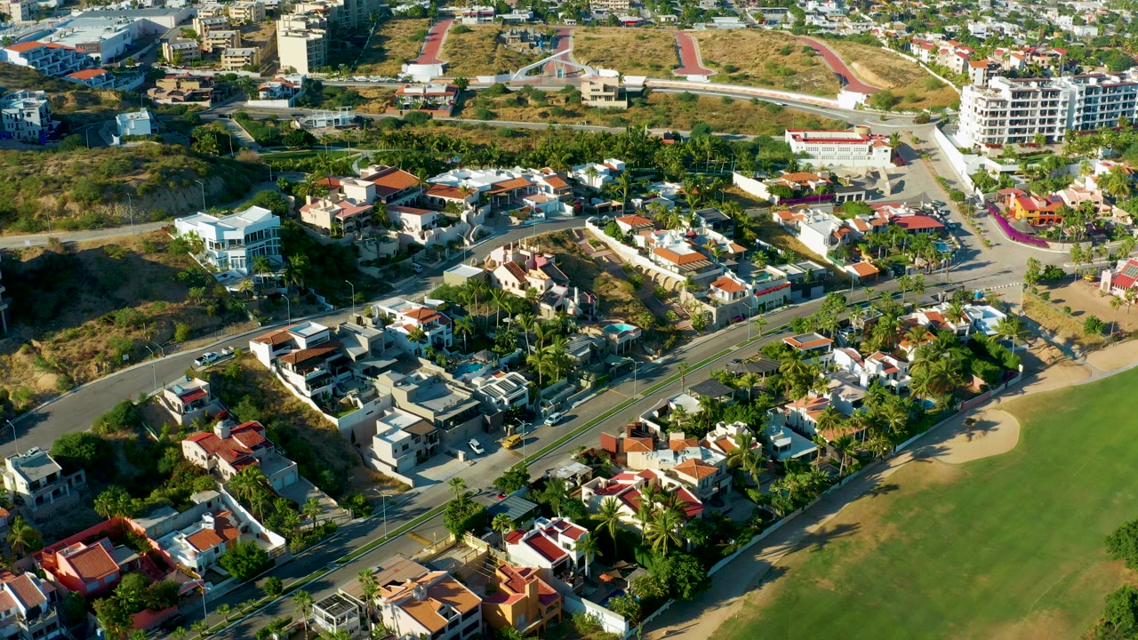 Aerial view over San Jose Del Cabo homes with red roofs, palm trees, a curving road, green grass, sandy ground, and no visible people.