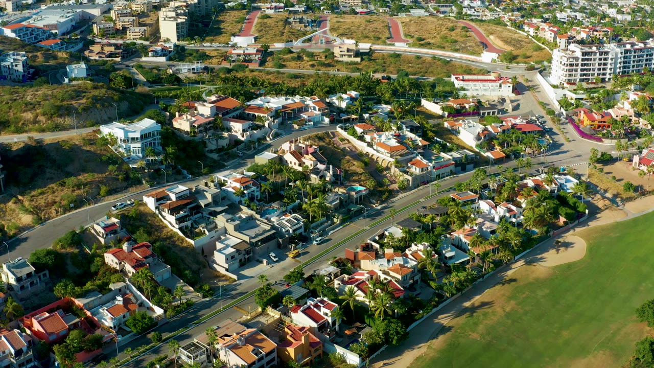 San Jose Del Cabo aerial frame showing houses, palm-lined lots, a paved road, open green ground at right, hillside buildings, and no visible people.
