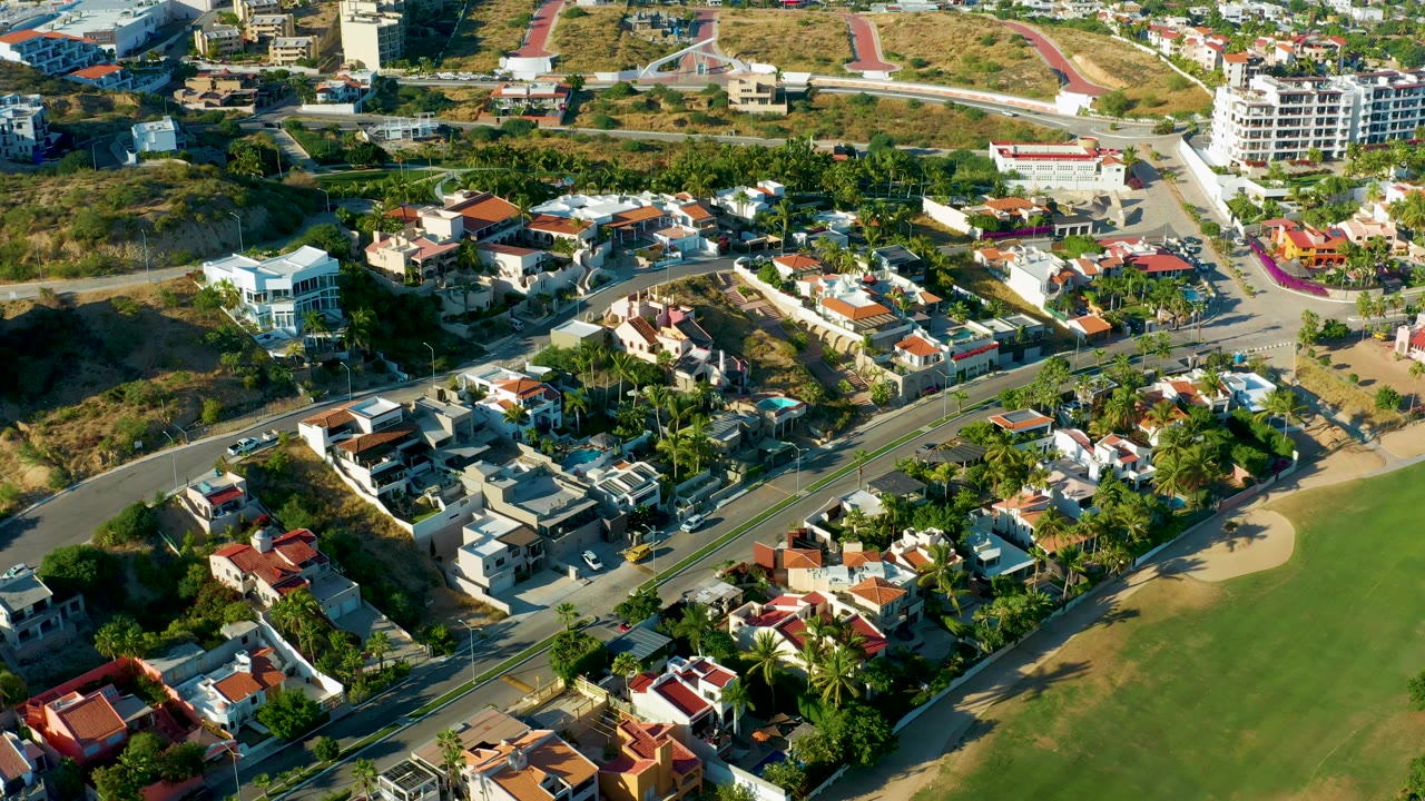 High aerial view in San Jose Del Cabo with red and white residential roofs, palms, intersecting roads, scrubby hillside, and no visible people.
