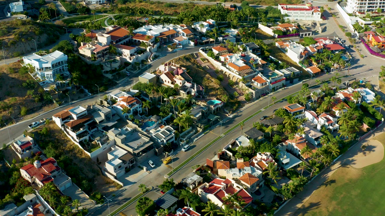 Oblique aerial view over San Jose Del Cabo homes, palm trees, dry hillside lots, and a curved road with small cars visible, with no visible people.
