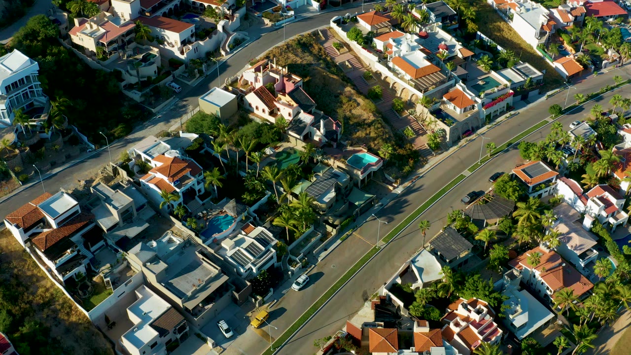 Aerial view of San Jose Del Cabo residential blocks, showing the central roadway, light-colored villas, red tile roofs, pool courtyards, trees, cars, and no visible people.