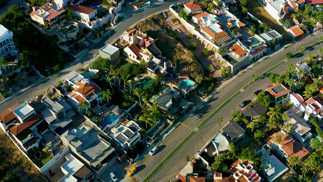 High oblique frame over San Jose Del Cabo houses and a divided road, with palm-lined lots, parked cars, sunlit rooftops, shaded yards, and no visible people.