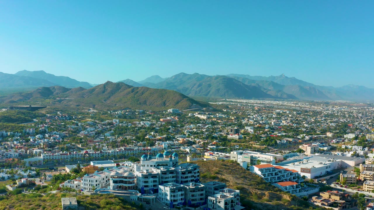 Opening frame of San Jose del Cabo from above, showing hillside residences in front, dense city blocks beyond, mountain ridges on the horizon, clear blue sky, and no visible people.