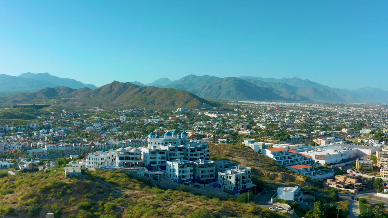 Aerial frame in San Jose del Cabo showing clustered hillside residences, a dense urban plain beyond, hazy mountain layers, clear daylight, and no visible people.