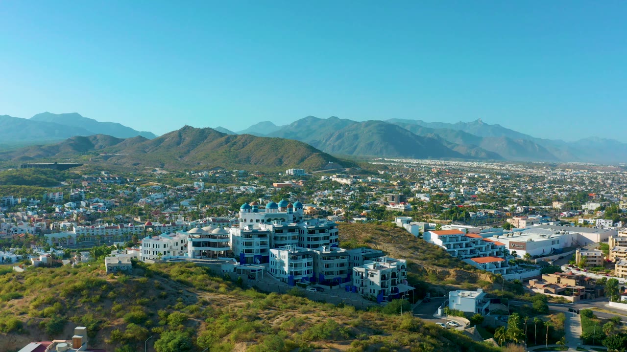 Drone view across hillside buildings toward a city and mountain range.