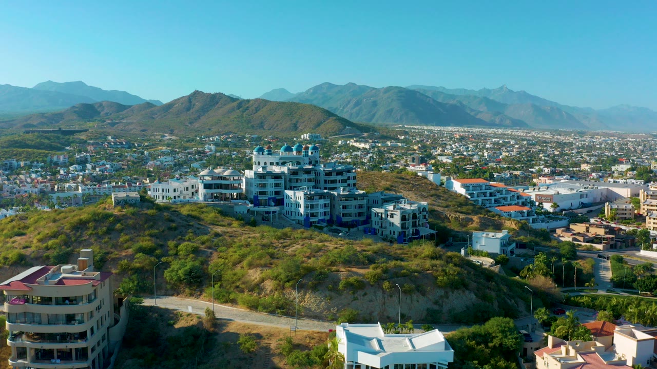 Wide aerial view in San Jose Del Cabo showing hillside residences, a winding road, distant city blocks, layered mountains, and no visible people.