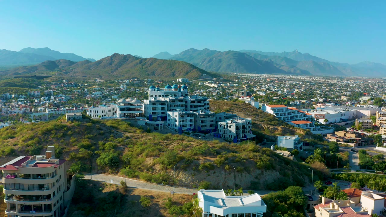 Aerial frame in San Jose Del Cabo with white and blue residential buildings on a green hillside, a road below, city development beyond, and no visible people.