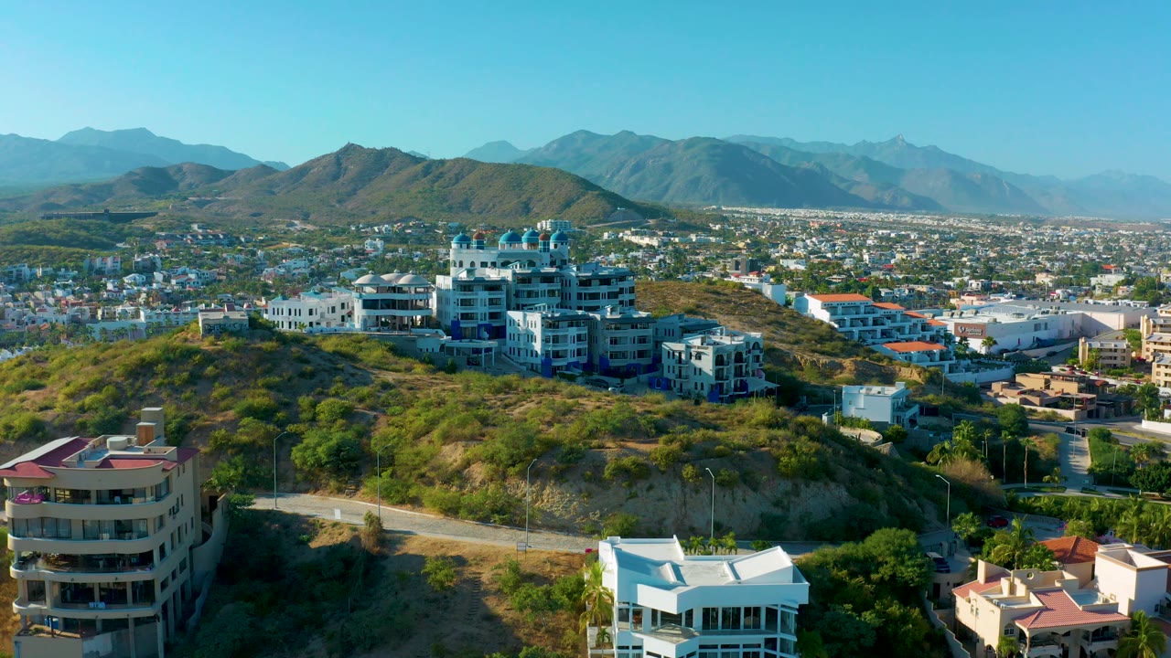 San Jose Del Cabo aerial view with a large residential building near the hill crest, foreground homes, dry green slopes, city sprawl, and no visible people.