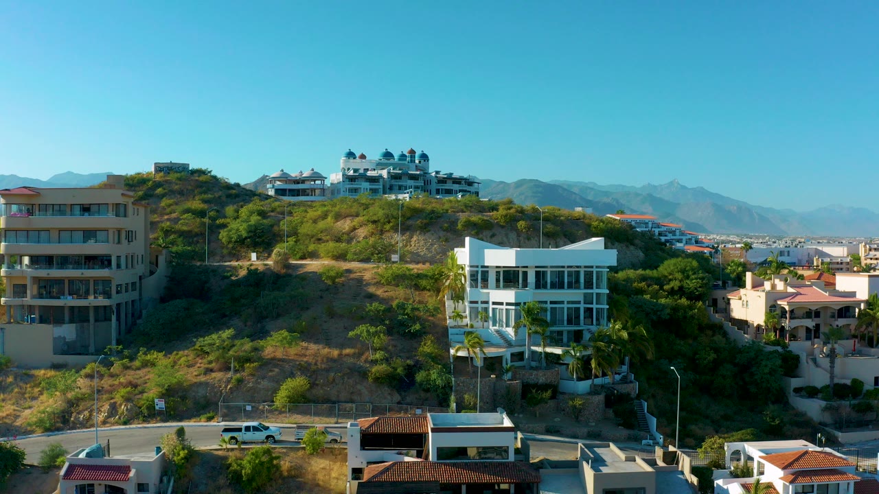 Wide aerial frame of San Jose Del Cabo hillside residences, with a beige apartment building at left, a white modern house at right, distant mountains, and no visible people.