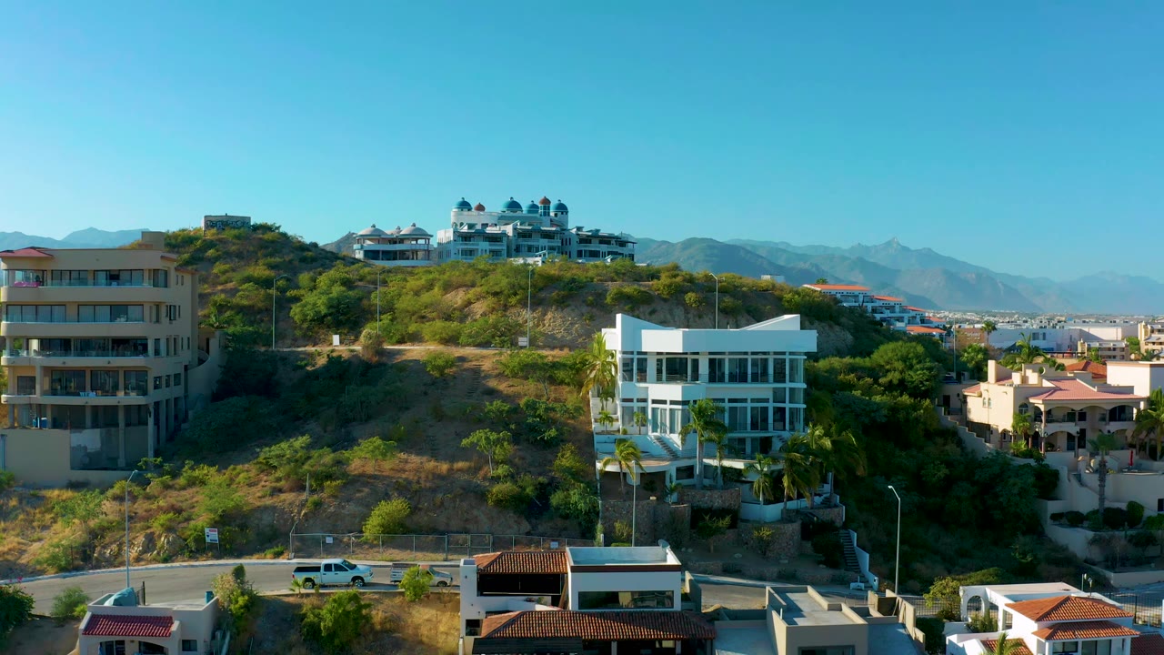 San Jose Del Cabo hillside neighborhood seen from above, with the white car more noticeable on the foreground road and layered homes climbing toward the ridge, with no visible people.