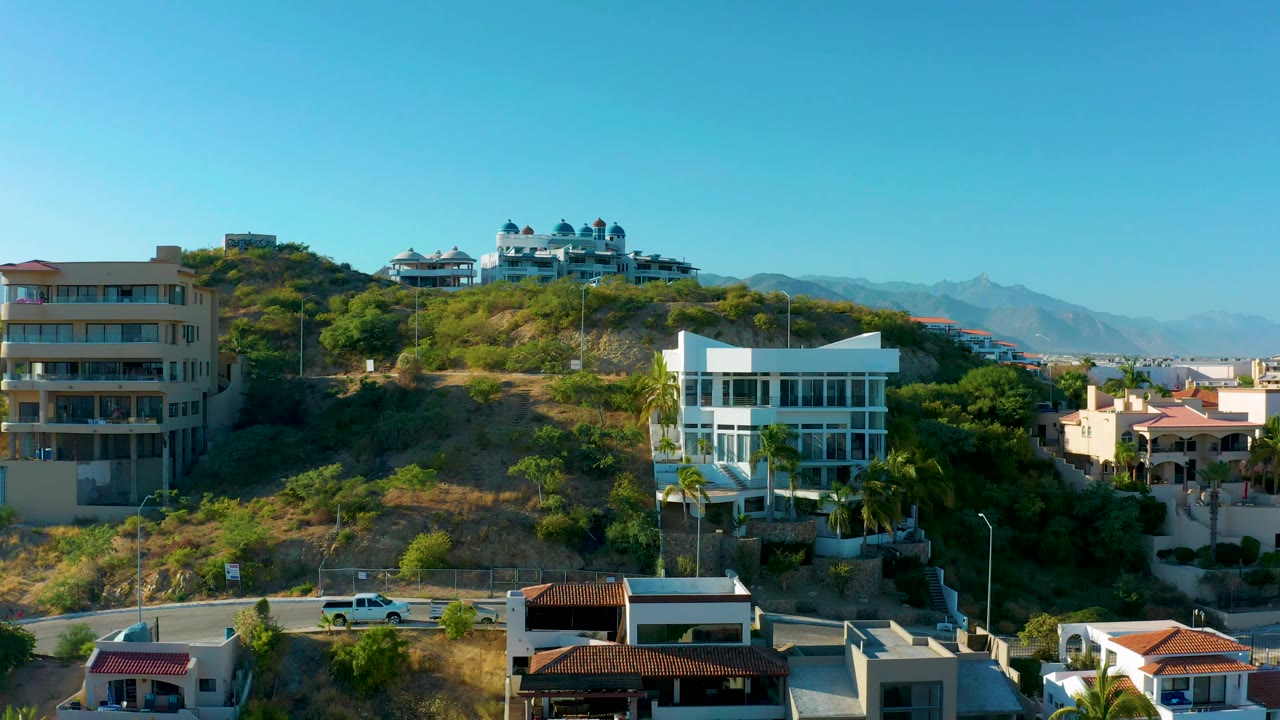 Aerial view in San Jose Del Cabo with a modern white house on the right, a beige multi-story building on the left, terraced hillside vegetation, and no visible people.