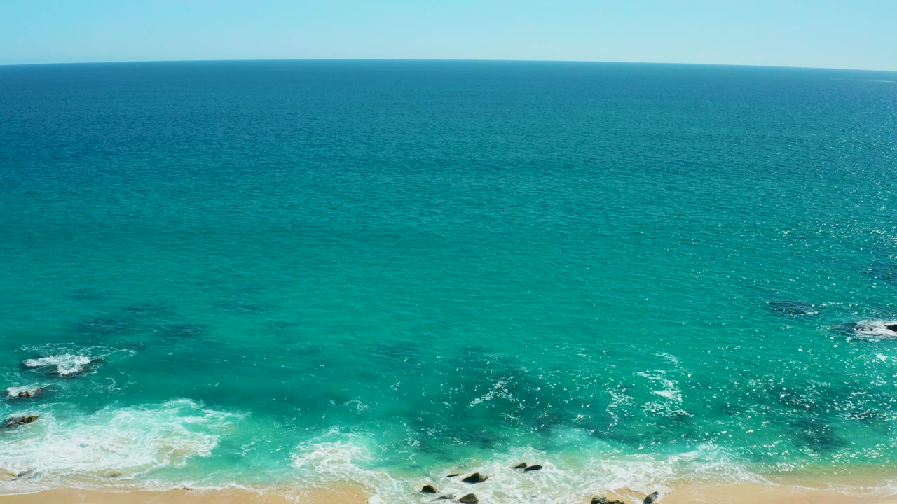 Aerial view of turquoise surf breaking on a sandy rocky shore.