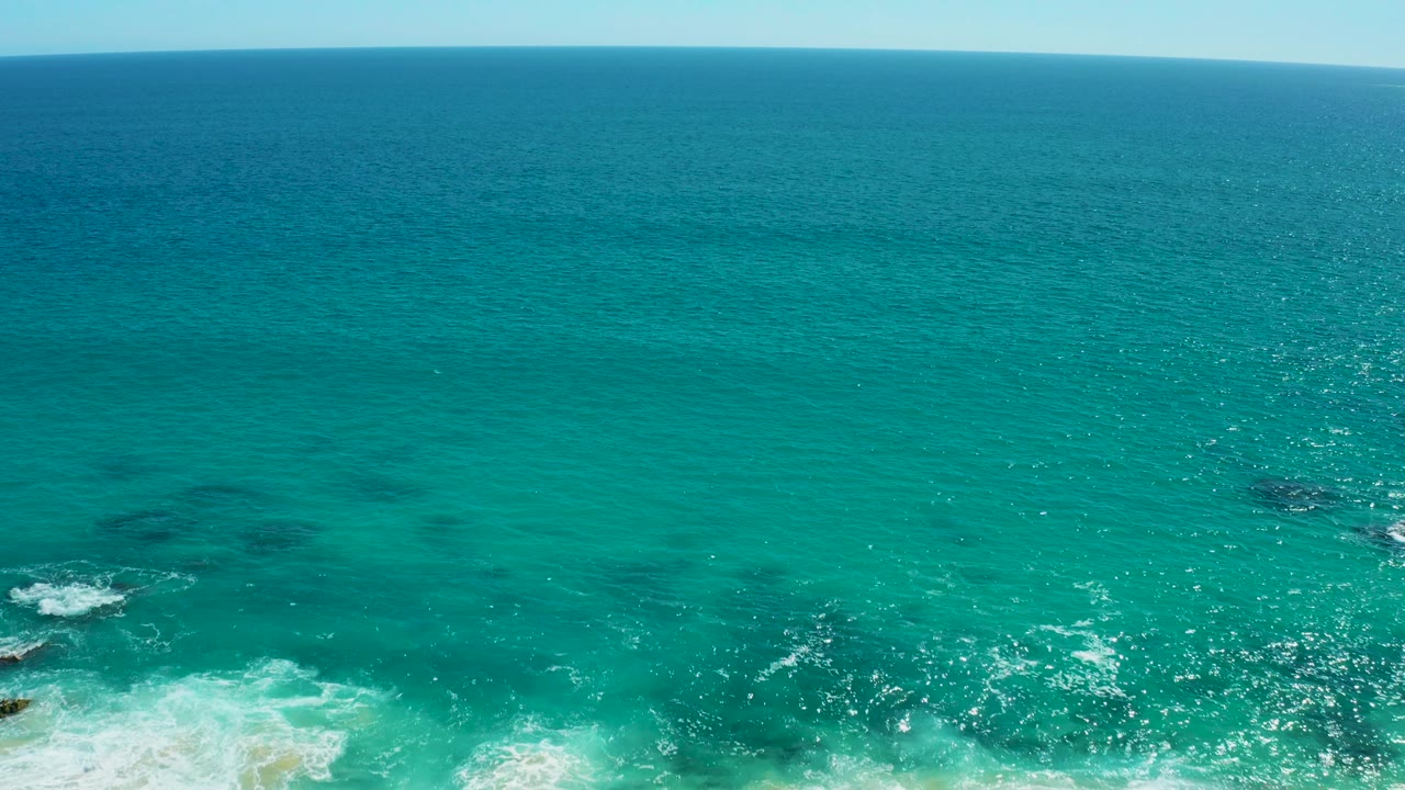 Aerial frame over East Cape BCS Mexico showing whitewater spread across turquoise shallows, small rocks near the beach, open sea beyond, and no visible people.