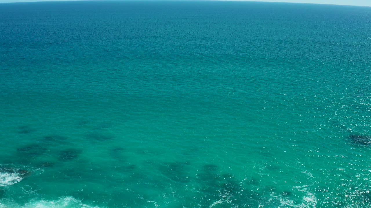 East Cape BCS Mexico ocean view with the beach nearly leaving frame, white surf at the lower edge, darker underwater areas, sunlit turquoise water, and no visible people.