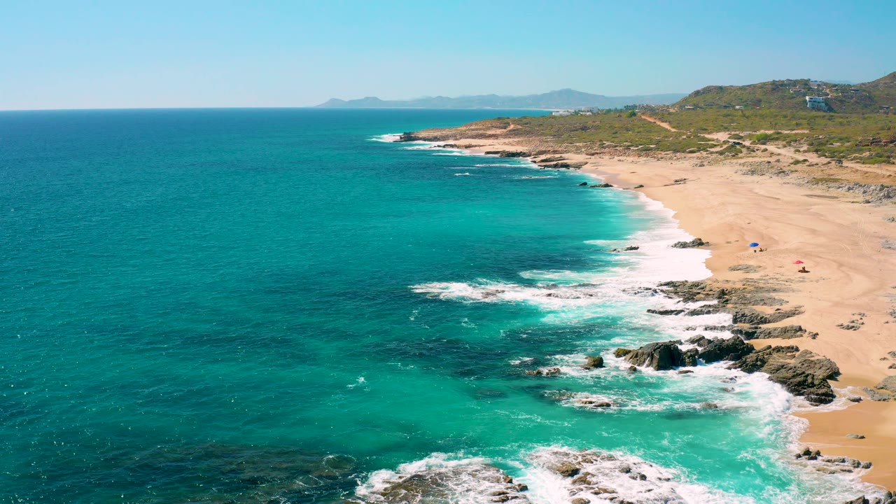 Aerial view of East Cape BCS Mexico with turquoise water on the left, white surf around dark rocks below, sandy beach to the right, distant hills on the horizon, and no visible people.