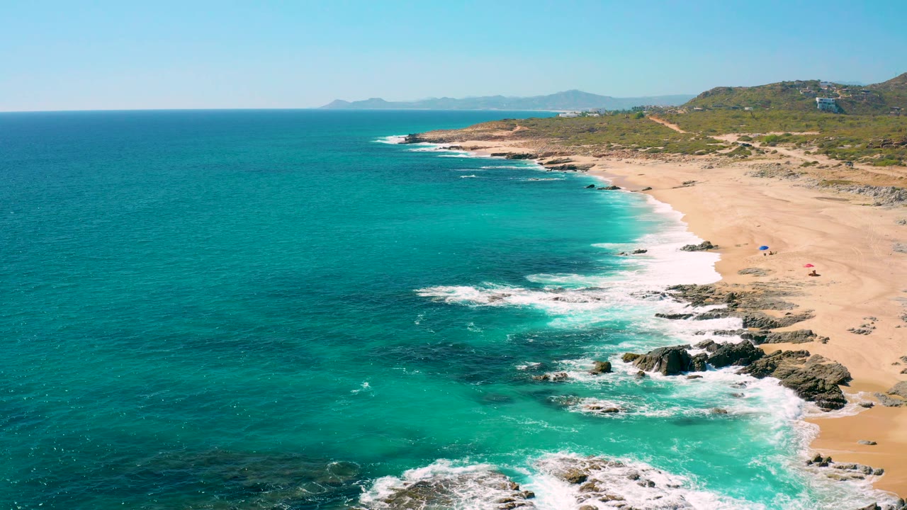Turquoise ocean curves along a sandy shore with waves breaking over rocks.