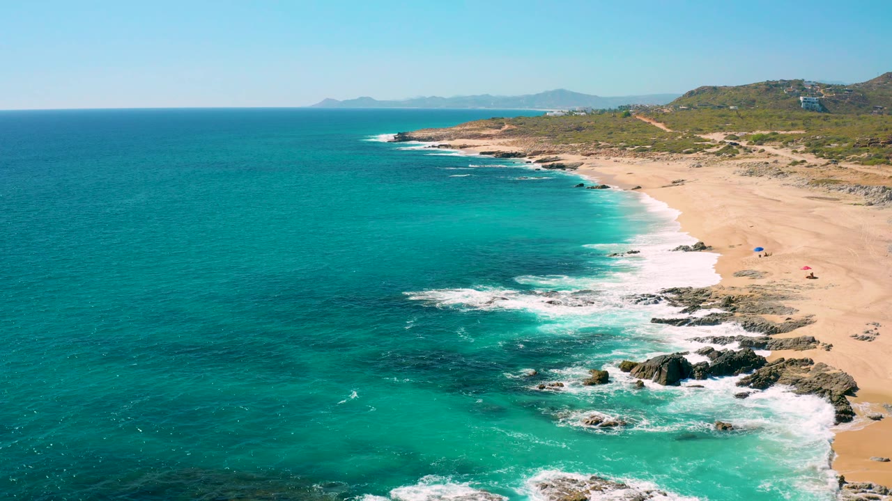 East Cape BCS Mexico coastline from above, with blue ocean across the left, pale sand and small beach umbrellas on the right, surf breaking over rocks, hills inland, and no discernible people.