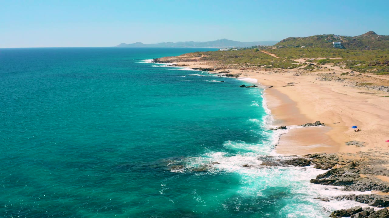 Aerial view over East Cape coastline with the surf line curving along the sandy beach, rocks in the lower right, deep turquoise water offshore, and no visible people.