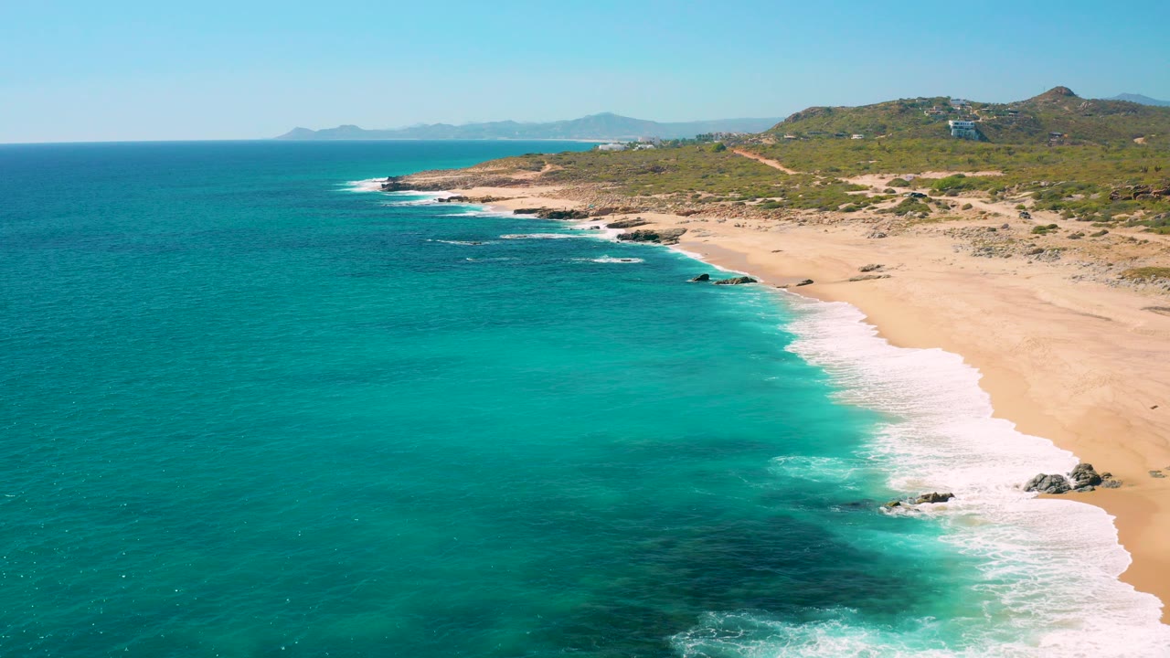 Aerial frame over East Cape Baja California Sur with turquoise water in the foreground, white surf on a sandy beach, rocky shoreline, green hills, distant landforms, and no visible people.