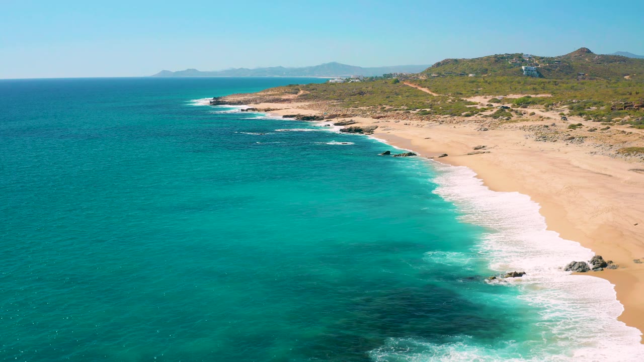 Aerial view of East Cape Baja California Sur with more open turquoise water filling the lower frame, a diagonal band of surf, sandy beach to the right, rocky points ahead, and no visible people.