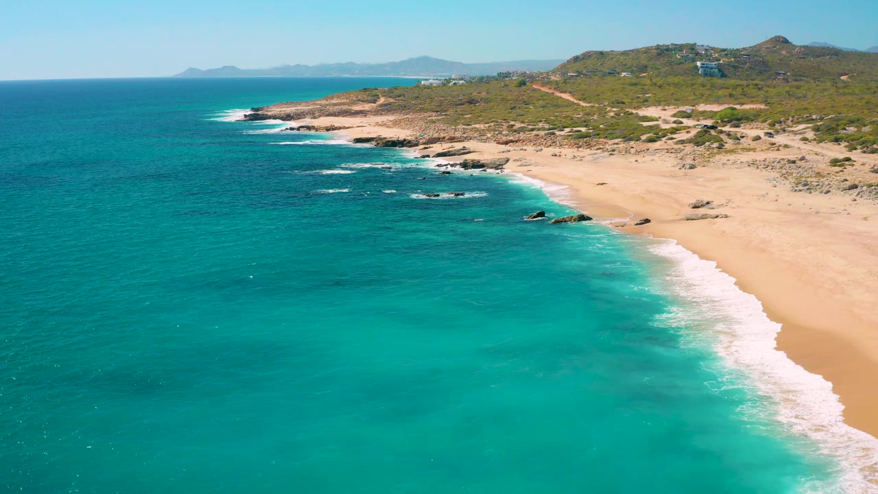 Aerial view in East Cape, Baja California Sur showing white foam tracing the sandy shore, dark rocks near the surf line, green hills behind the beach, and no visible people.