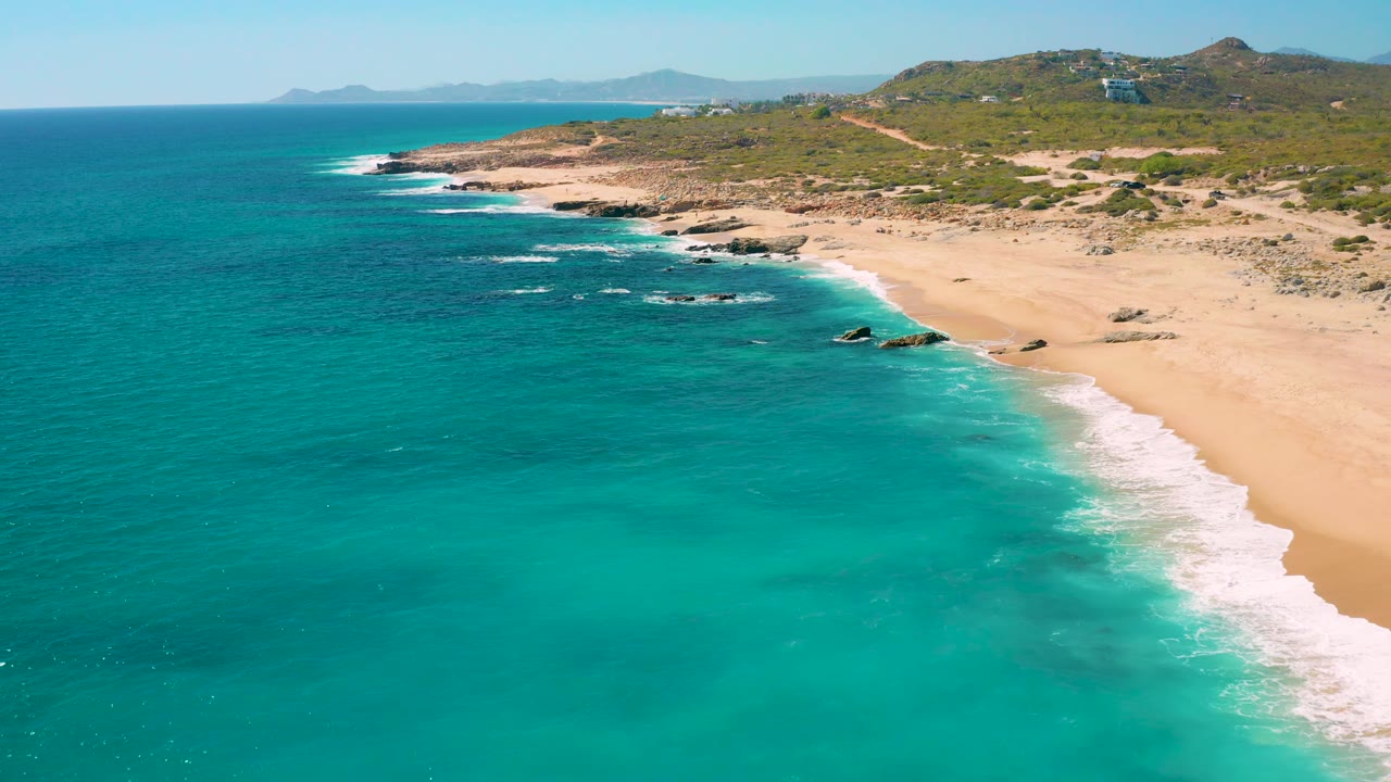 Aerial frame over East Cape, Baja California Sur with more open turquoise water in front, a bright strip of surf at right, rocky points along the coast, and no visible people.