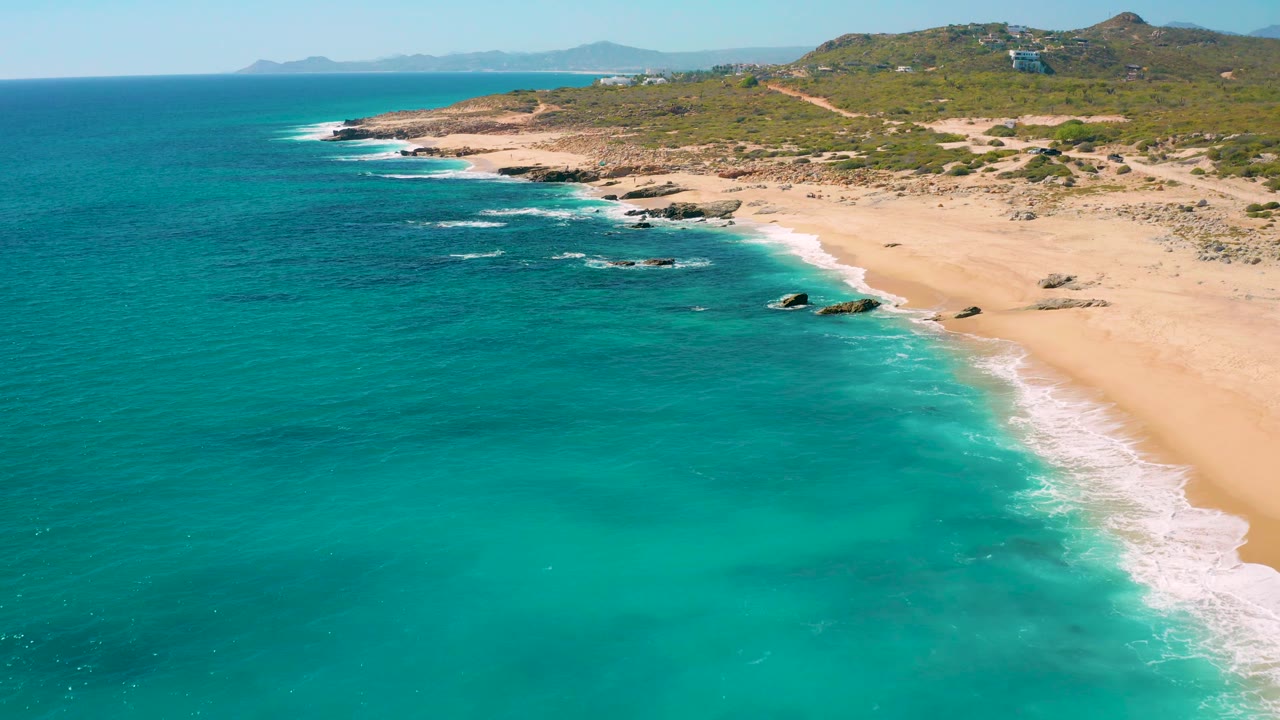 Aerial coastline with turquoise sea, sandy beach, rocks, and hills.