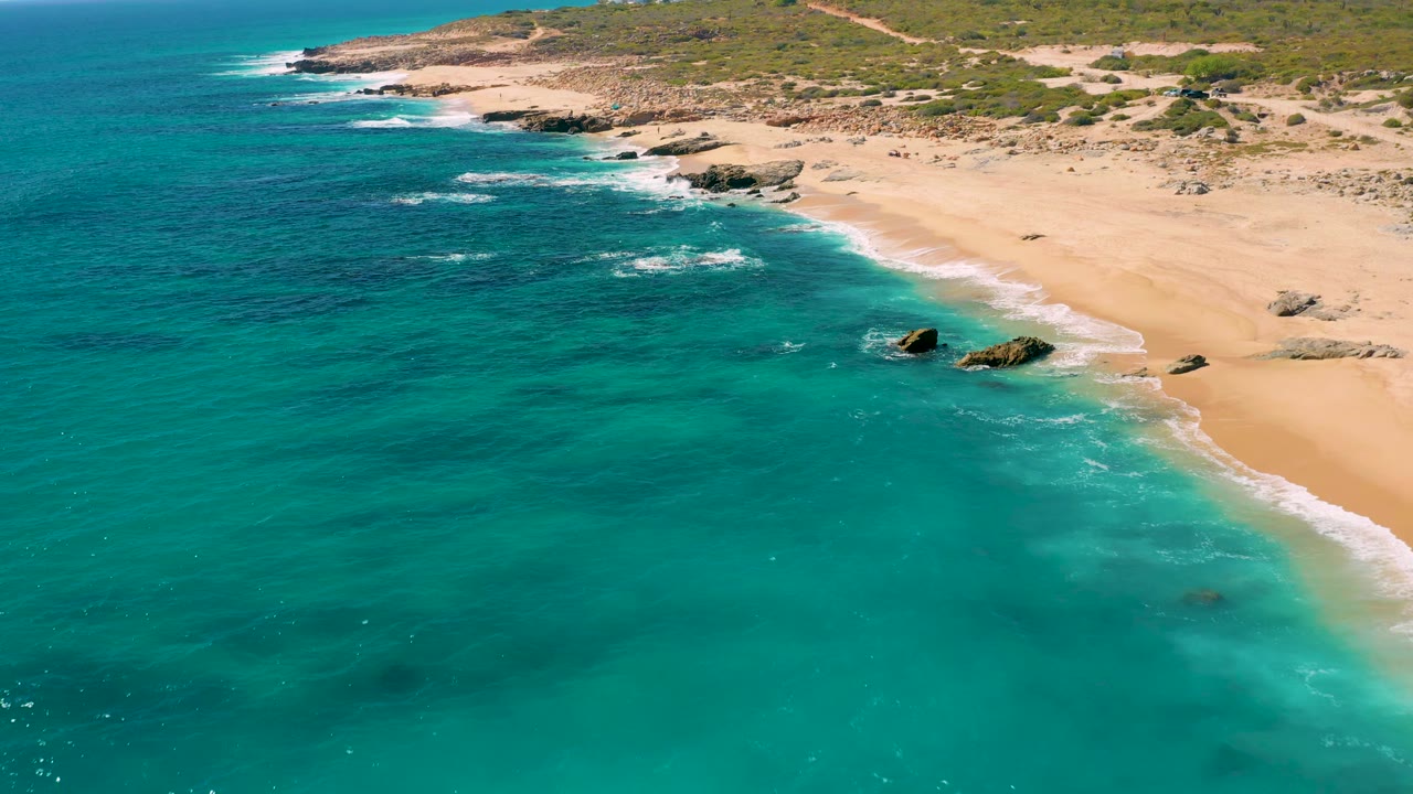 East Cape, Baja California Sur shoreline seen from above, with turquoise water in the foreground, white foam along the sand, and no visible people.