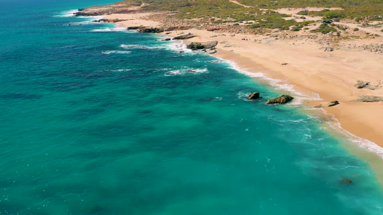 Aerial East Cape coastline frame with blue-green water covering most of the view, a sandy beach at right, scattered rocks, and no visible people.