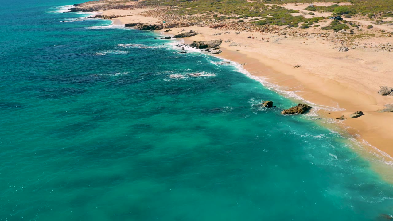 East Cape, Baja California Sur beach from an aerial angle, showing turquoise shallows, pale sand, rocks close to the waterline, and no visible people.