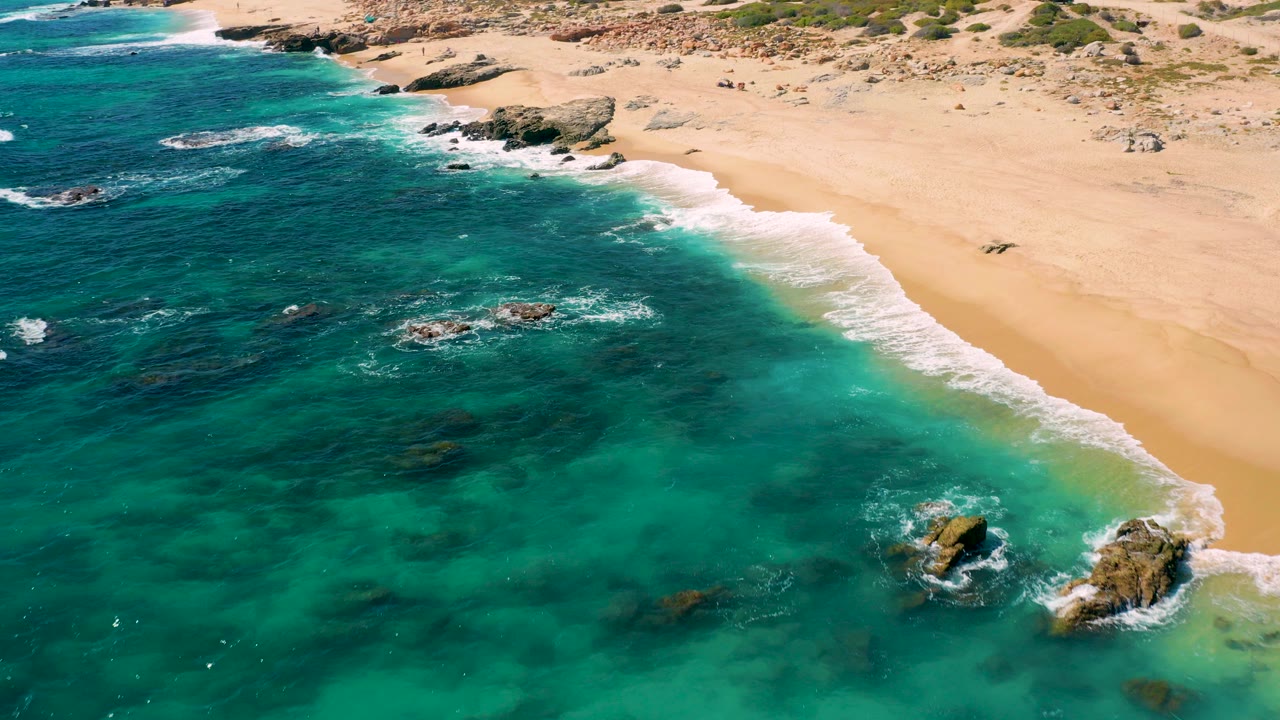 Aerial frame over East Cape, Baja California Sur, with turquoise ocean on the left, white surf tracing the sand, rocky outcrops near shore, and no visible people in this moment.