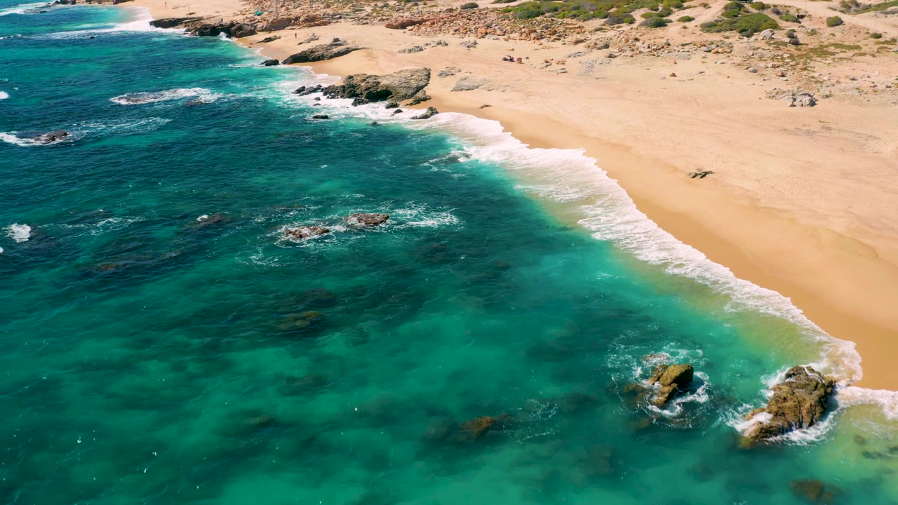 Turquoise water and foam lines meet a broad sandy beach with rocks offshore.