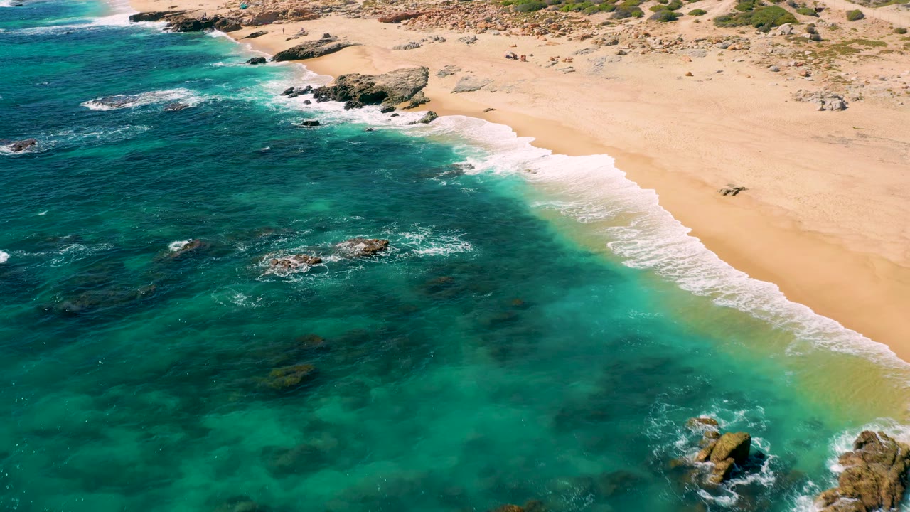 The East Cape beach remains in aerial view as rocks cluster near the surf zone, green-blue water fills the foreground, and the empty-looking sand stretches across the upper frame.