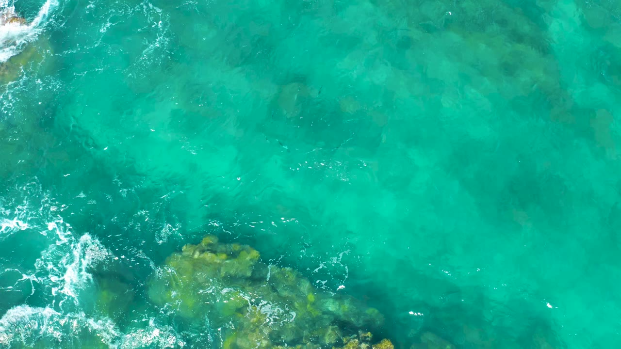 Overhead frame of East Cape turquoise water with submerged rocks near the lower edge, small white foam patches on the left, and no visible people.