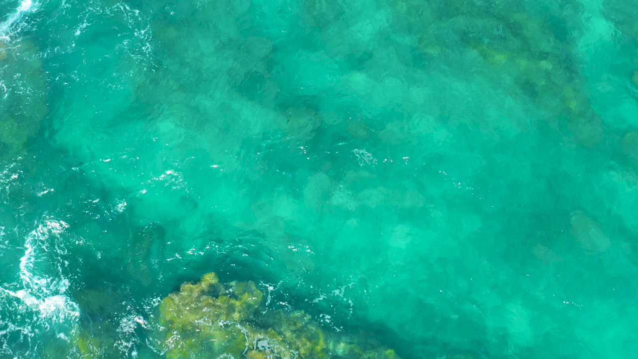 Aerial view of clear blue-green water in East Cape, Baja California Sur, with reef shapes under the surface and a small band of foam at left, no visible people.