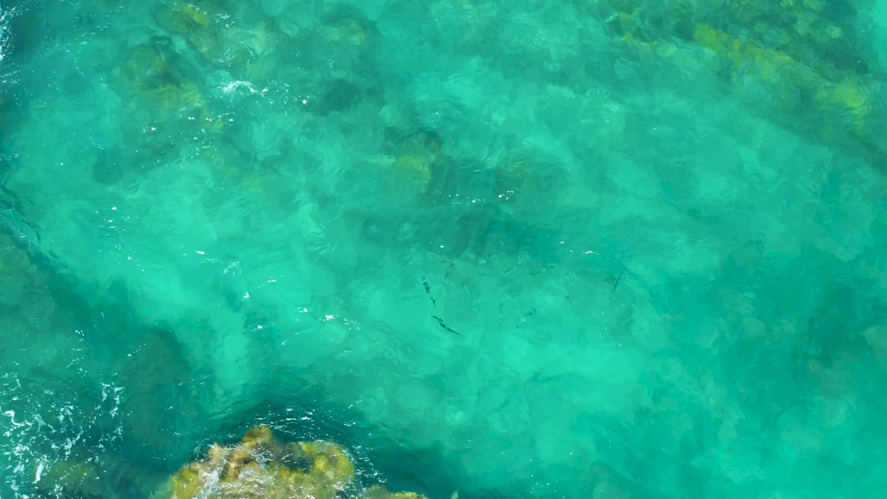 Aerial frame over East Cape BCS Mexico water with a larger submerged rock patch near the lower edge, pale ripples across the surface, and no visible people.
