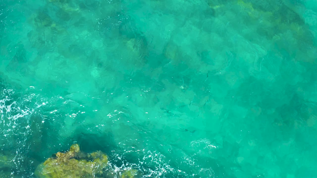 Clear East Cape ocean water seen from above, with a small foamy ripple crossing near the lower-left rocky patch and no visible people.